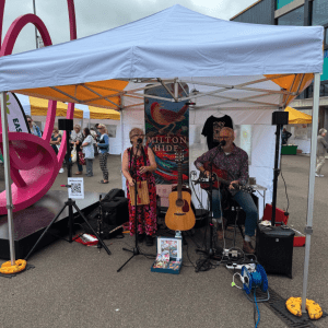 couple stand under gazebo playing guitar and singing in the middle of a market square with people milling around